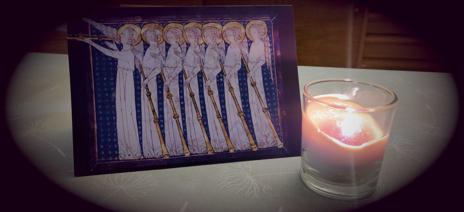 The Christmas card standing on a table by candlelight: the mediaeval illumination of seven haloed angels with long gold trumpets, on a deep blue ground, printed on a folded card, with a lit candle in a glass beside it.